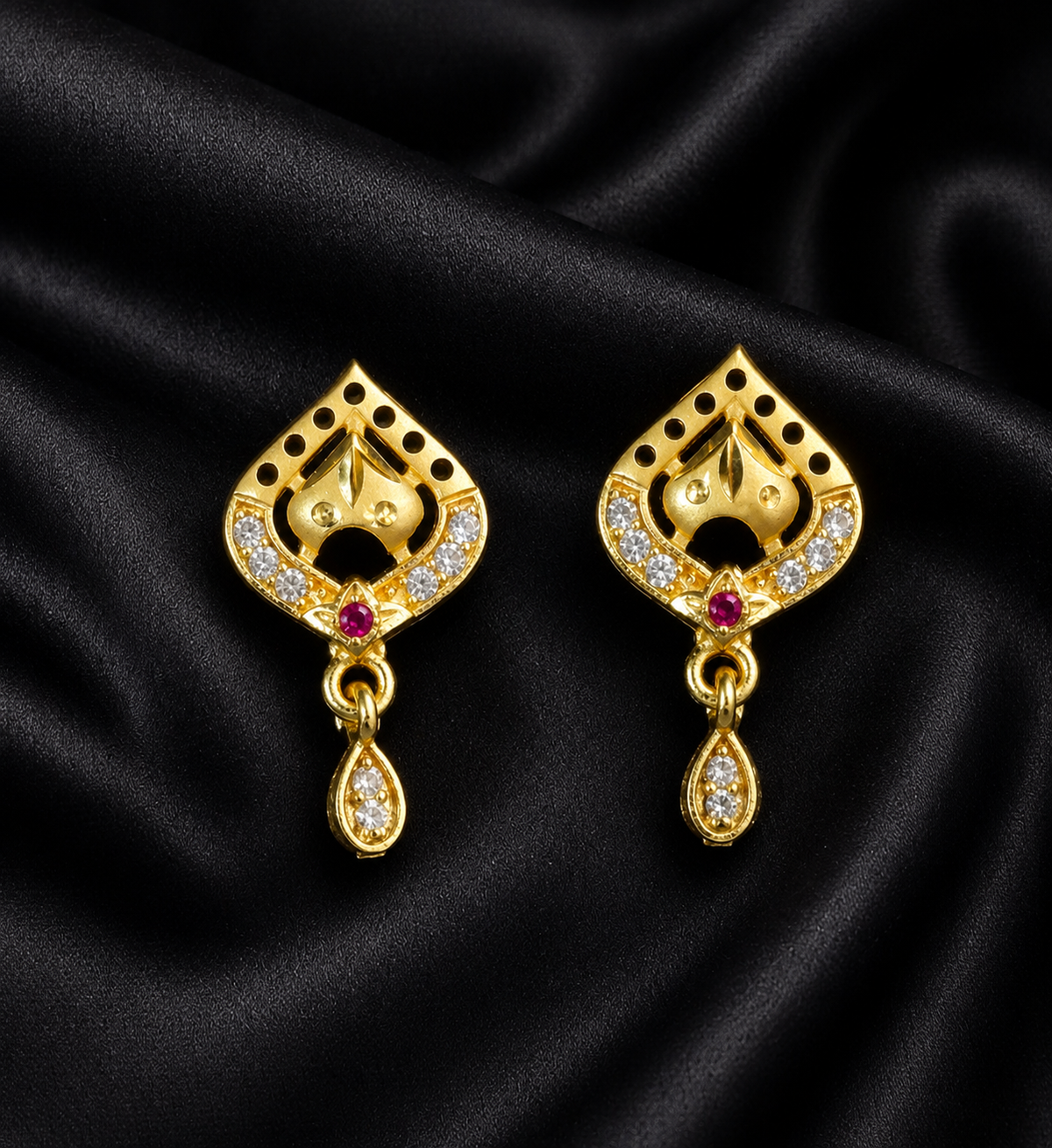 A pair of gold-toned traditional Indian earrings with intricate filigree cutouts, small white stones, and a tiny red gemstone, displayed on a black satin fabric background.