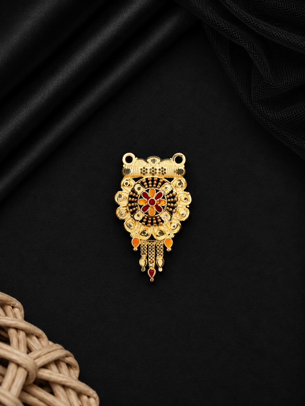 A gold-toned traditional Indian pendant with intricate filigree work, red and black enamel details, and hanging teardrop charms, displayed on a black satin fabric background.