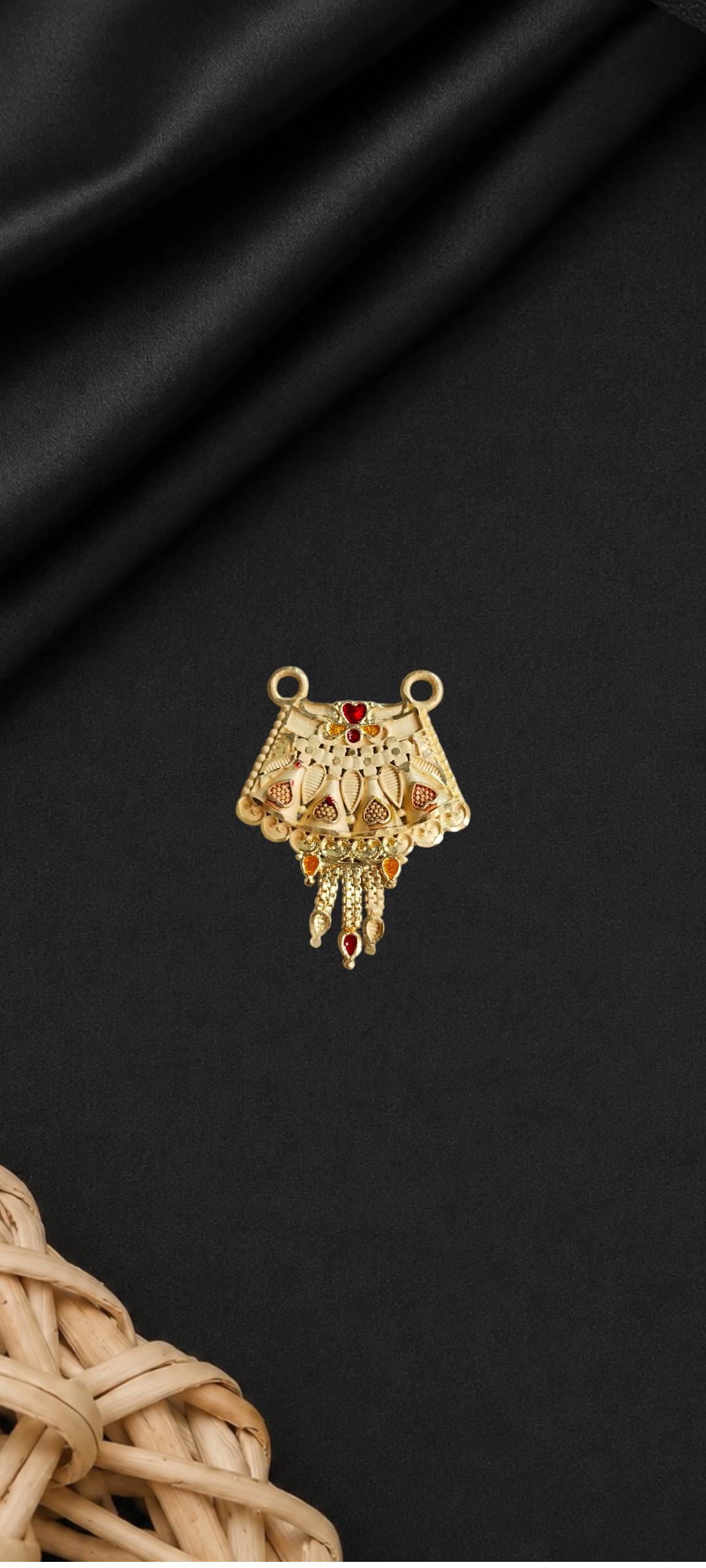 A gold-toned ornate jewelry charm or pendant with filigree detailing and small red gem accents, displayed on a dark satin fabric background with a woven wicker prop at the bottom left.