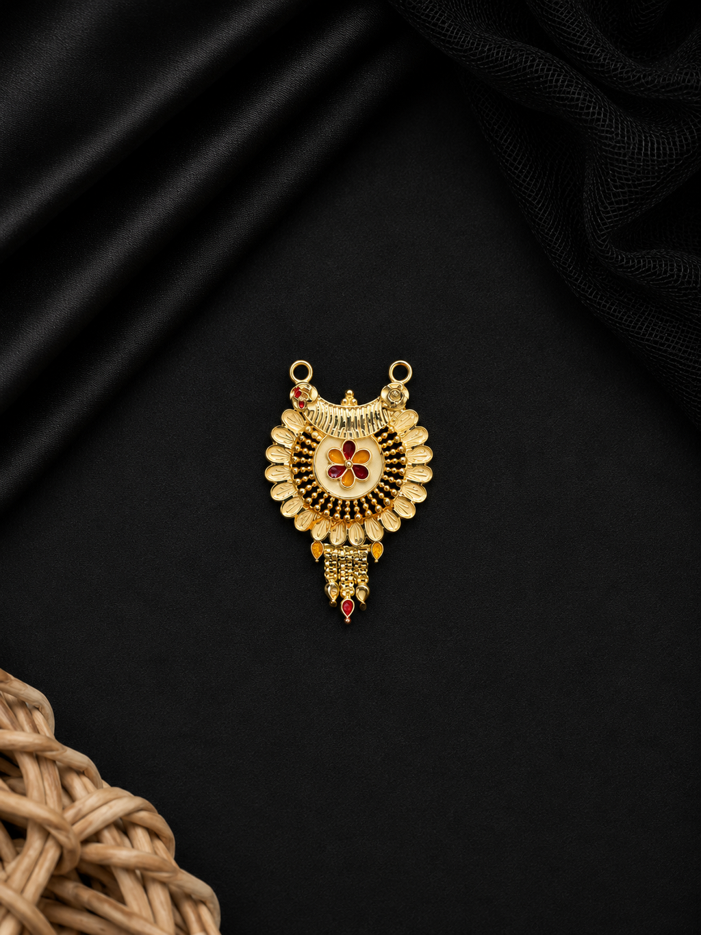 A gold-toned traditional pendant with intricate filigree, a central floral motif in red and gold, and small red gemstone accents, displayed on a black satin background with a woven straw prop in the corner.